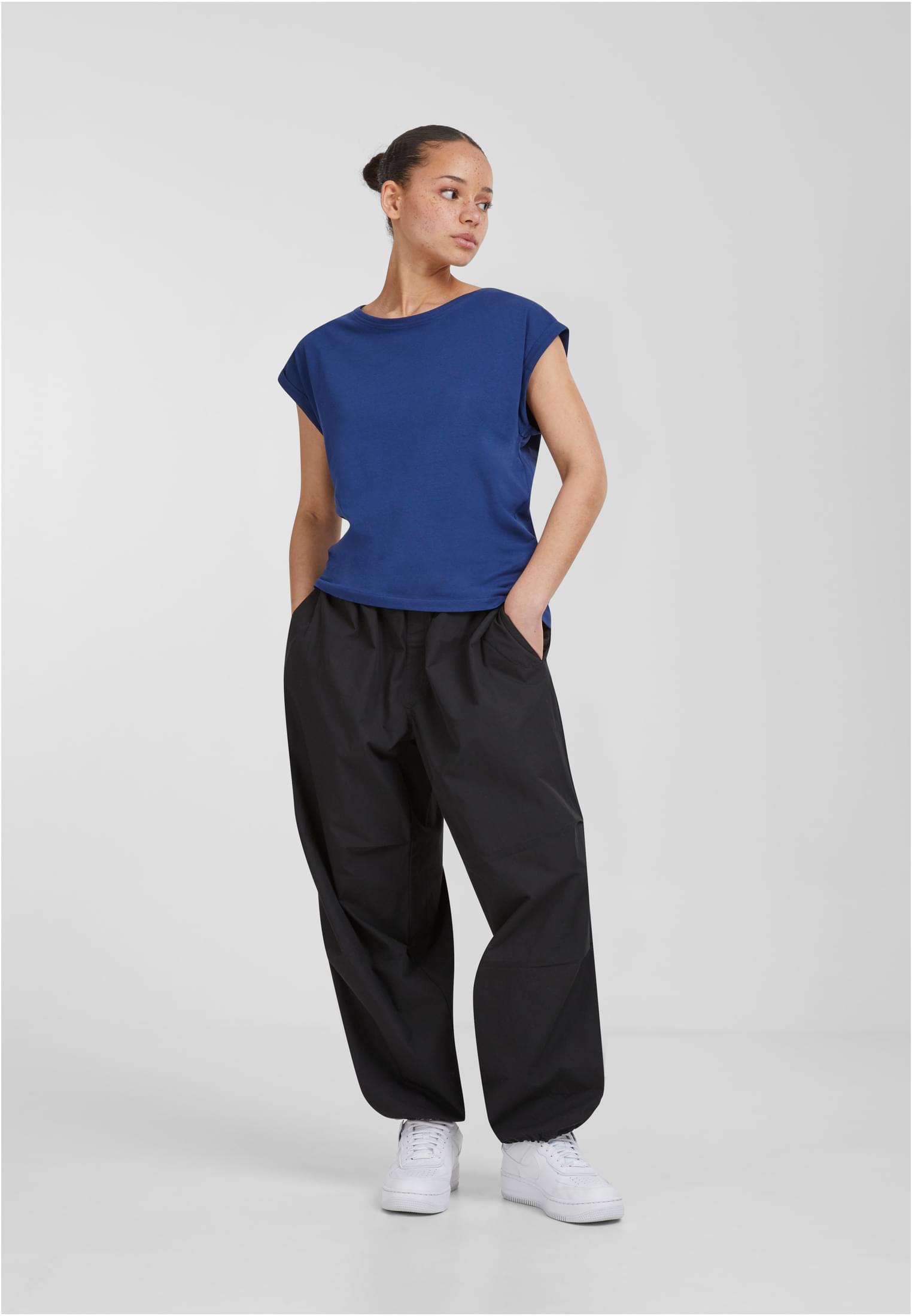 En kvinna som poserar i en blå Ladies Extended Shoulder Tee, svarta vida byxor och vita sneakers.