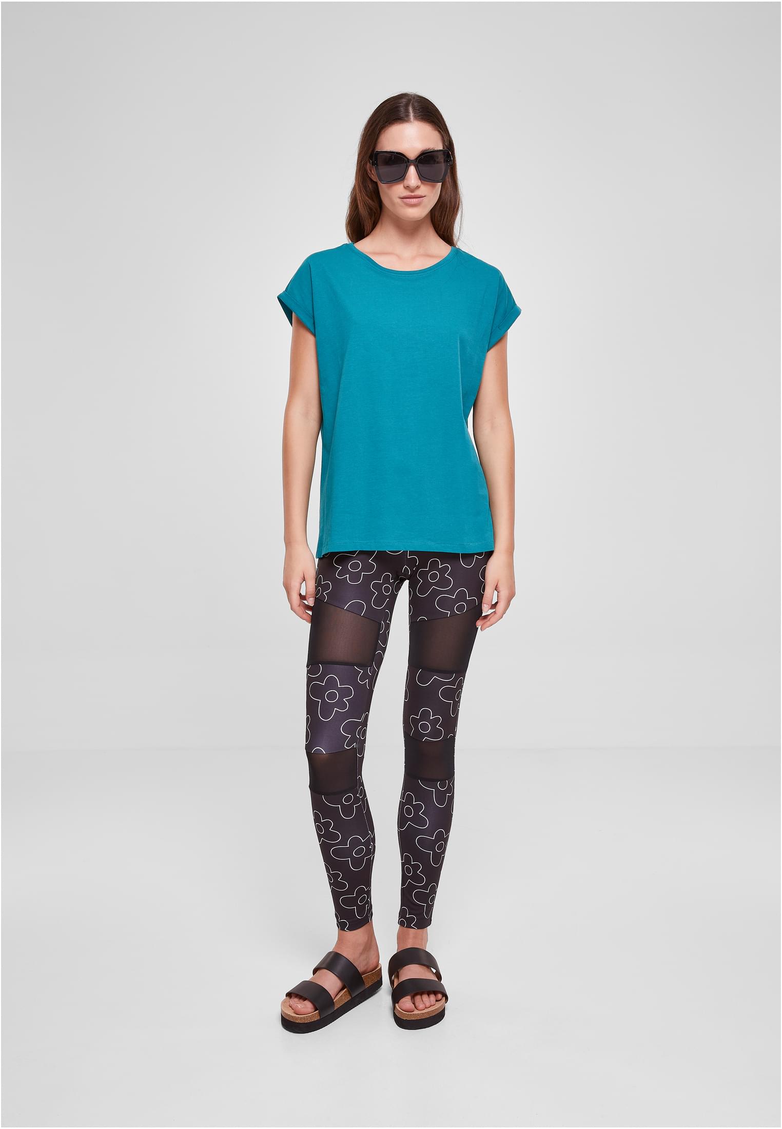 En kvinna visar framsidan av den turkosa t-shirten Ladies Extended Shoulder Tee, stylad med mönstrade leggings och solglasögon.