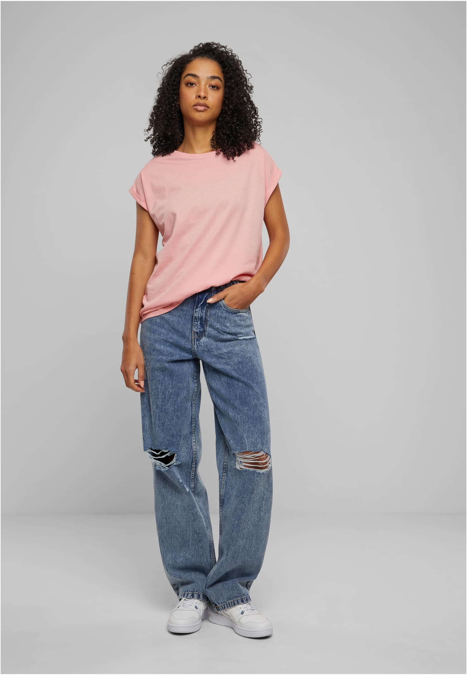 Kvinna med lockigt hår som bär den ljusrosa t-shirten Ladies Extended Shoulder Tee med slitna jeans och vita sneakers.
