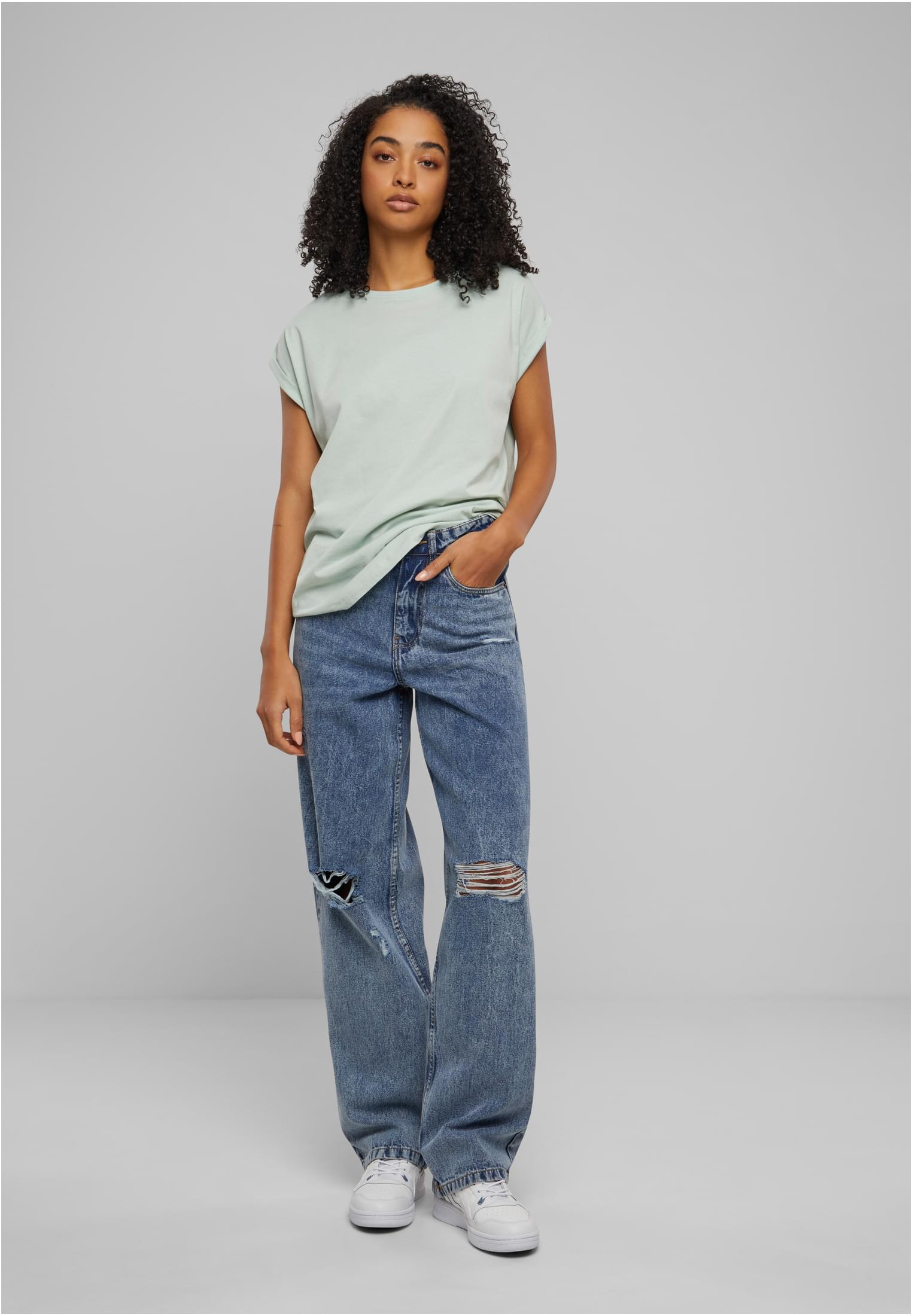 En kvinna i helfigur som bär en mintgrön t-shirt med nedhasade axlar och uppvikta ärmar, kombinerat med slitna blå jeans och vita sneakers.