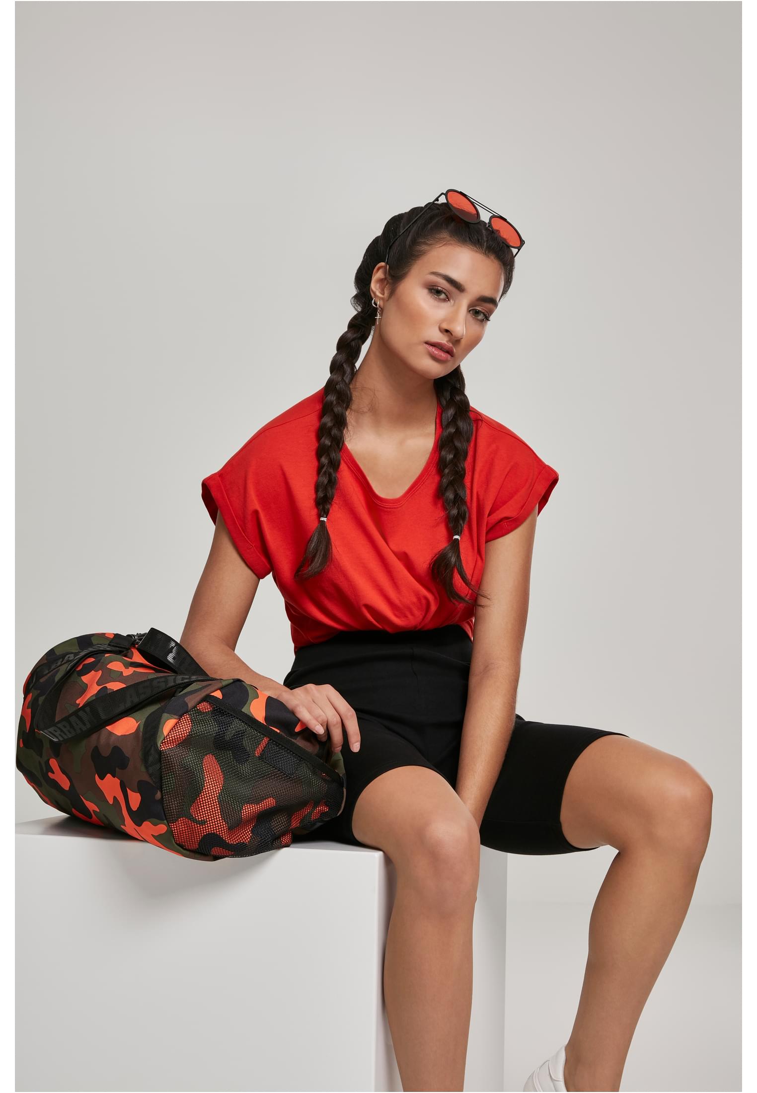 En kvinna poserar i en röd Ladies Extended Shoulder Tee från Urban Classics, stylad med svarta cykelbyxor och en kamouflagefärgad träningsväska.