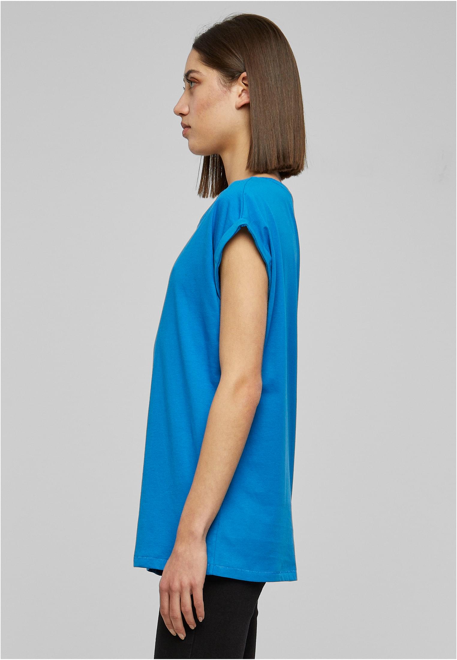 En kvinna fotograferad från sidan, klädd i en blå t-shirt med nedhasade axlar och uppvikta korta ärmar.