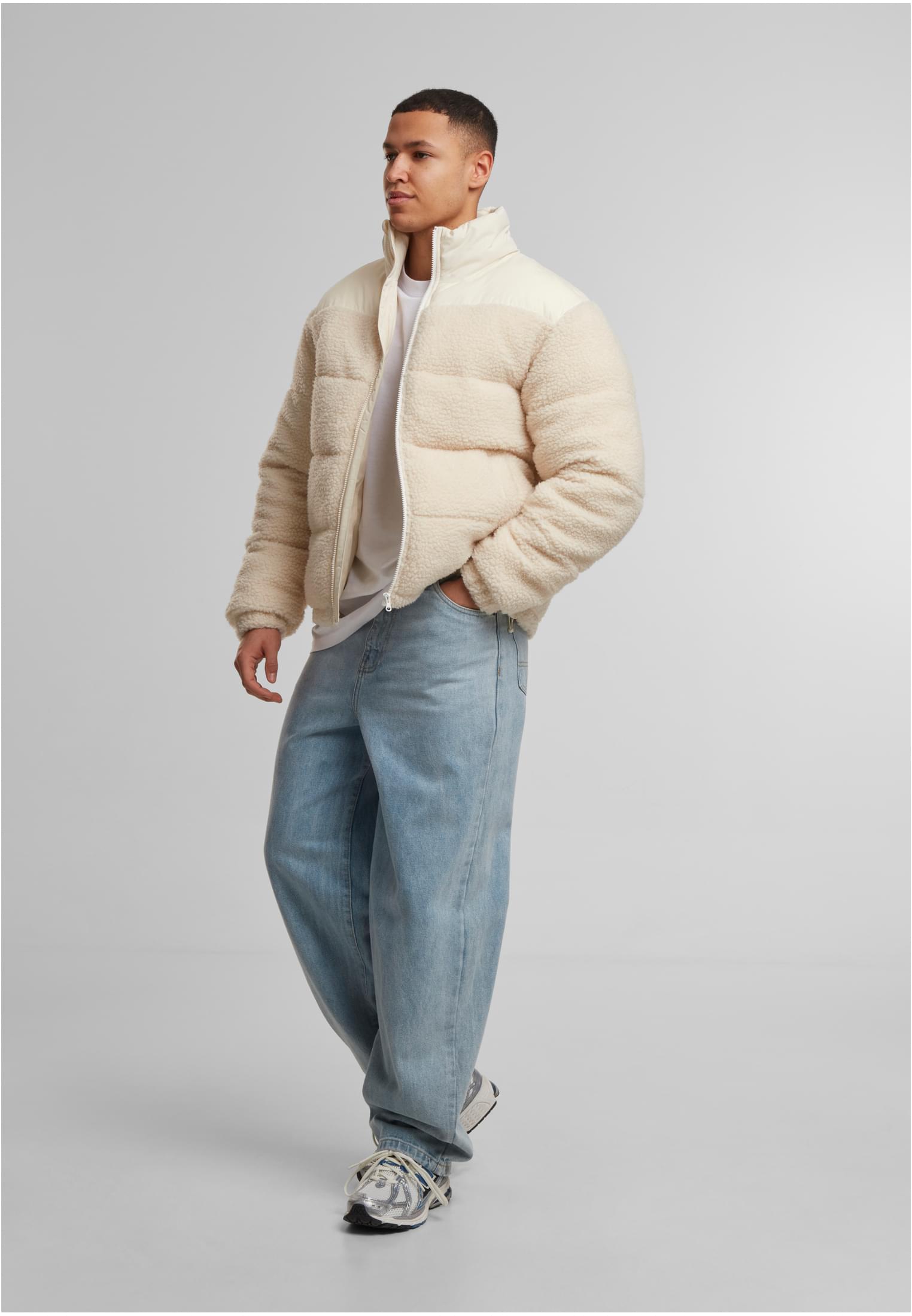 En man poserar i en beige teddy- och puffjacka, vit t-shirt och ljusa, vida jeans.