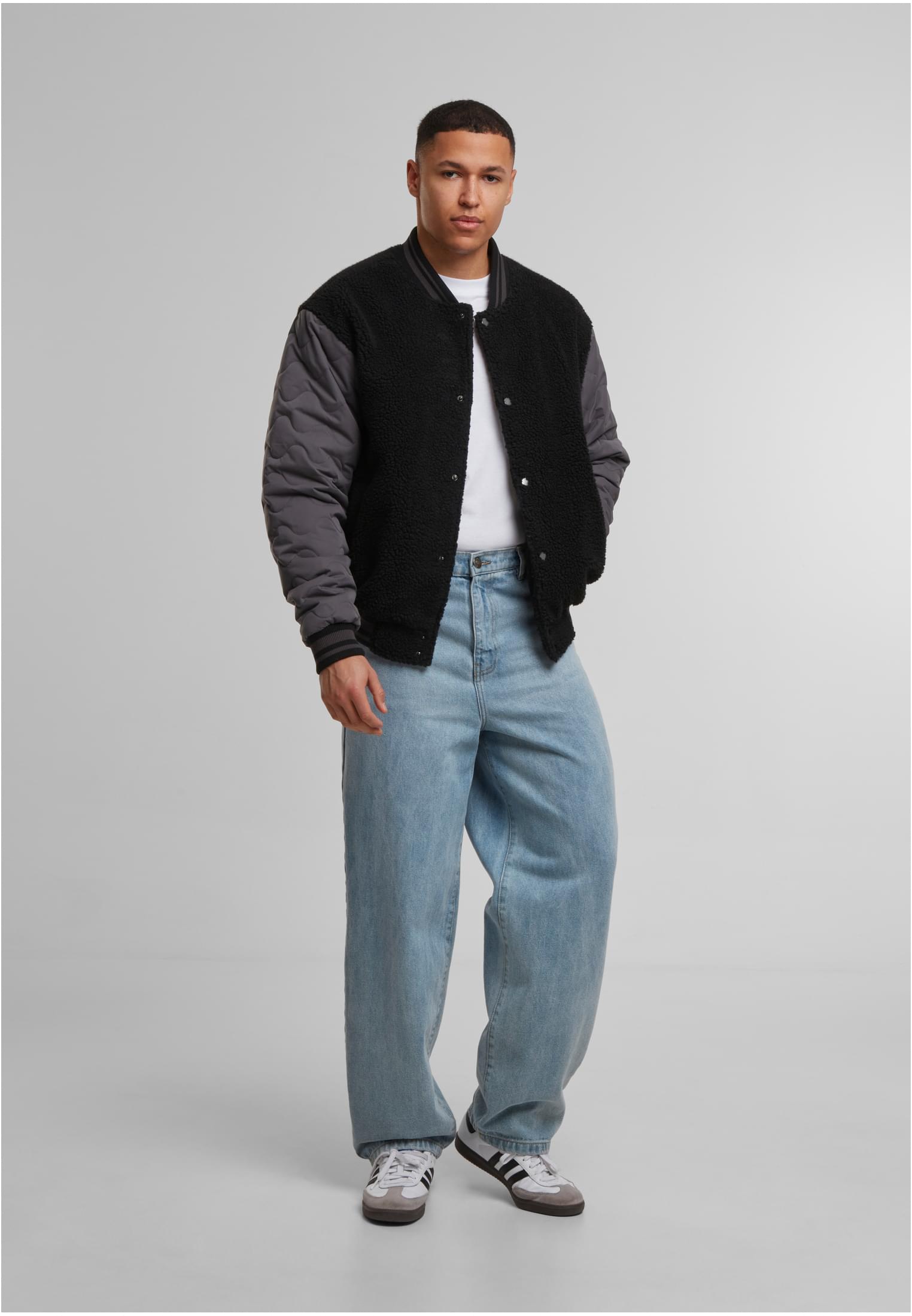 En man som bär en svart Teddy College-jacka med mörkgrå, kviltade ärmar, en vit t-shirt och ljusblå, vida jeans.