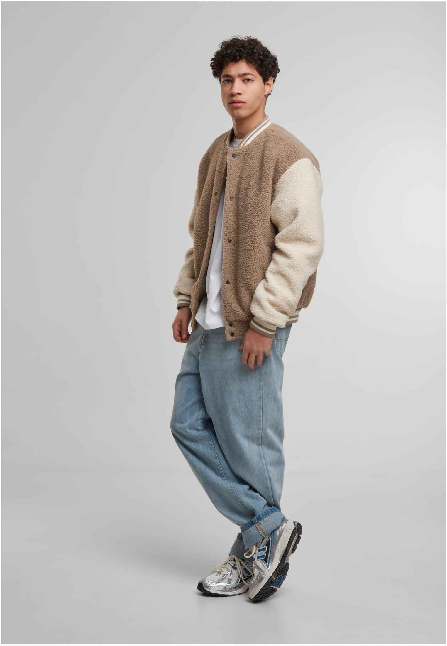 En manlig modell bär en Teddy College-jacka i darktaupe med benvita ärmar över en vit t-shirt och ljusblå jeans.