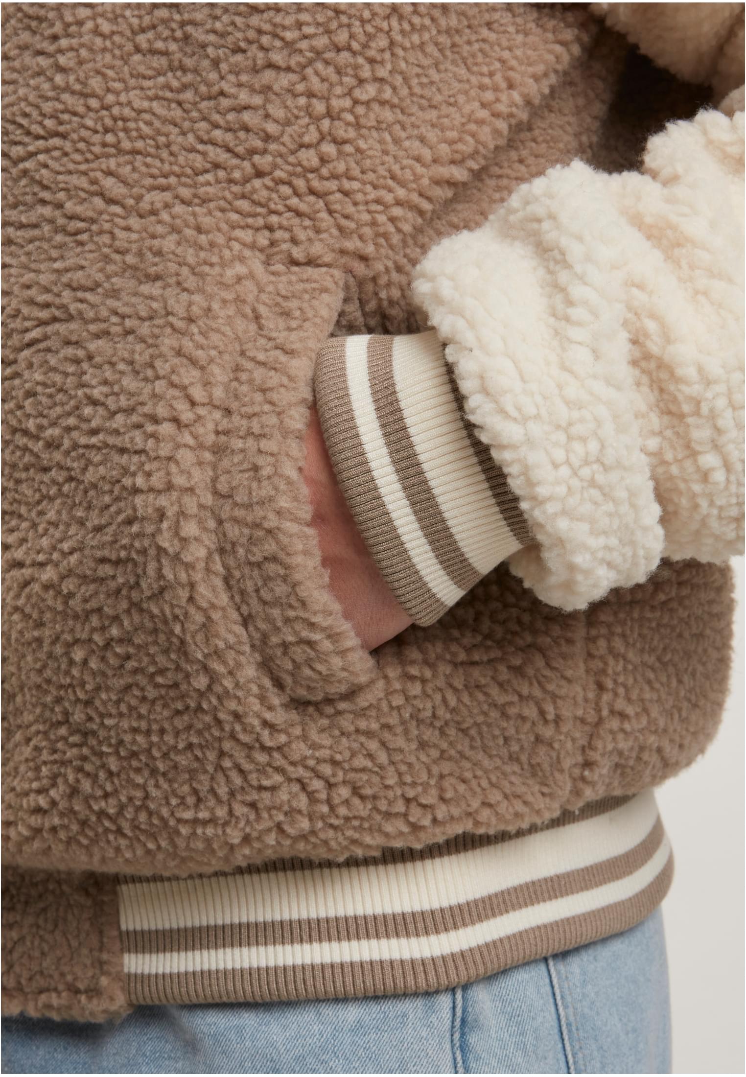 Närbild på en teddy-collegejacka i mörk taupe och benvitt, som visar tygets struktur, en hand i fickan och de randiga muddarna.