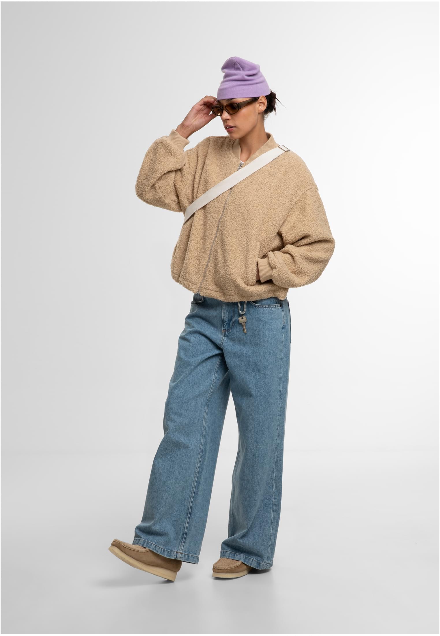 En kvinna som bär en beige UC Structured Bomber Jacket med vida jeans, lila mössa och solglasögon.