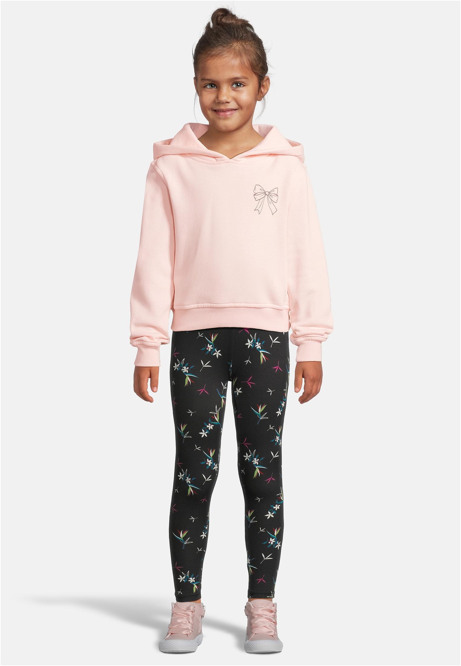 En flicka fotograferad framifrån klädd i en ljusrosa luvtröja med ett rosettryck, svarta leggings med blommönster och rosa skor.